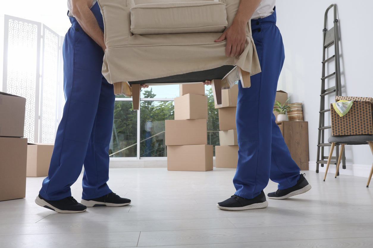 Two people in blue uniforms carrying a beige chair in a room with moving boxes.
